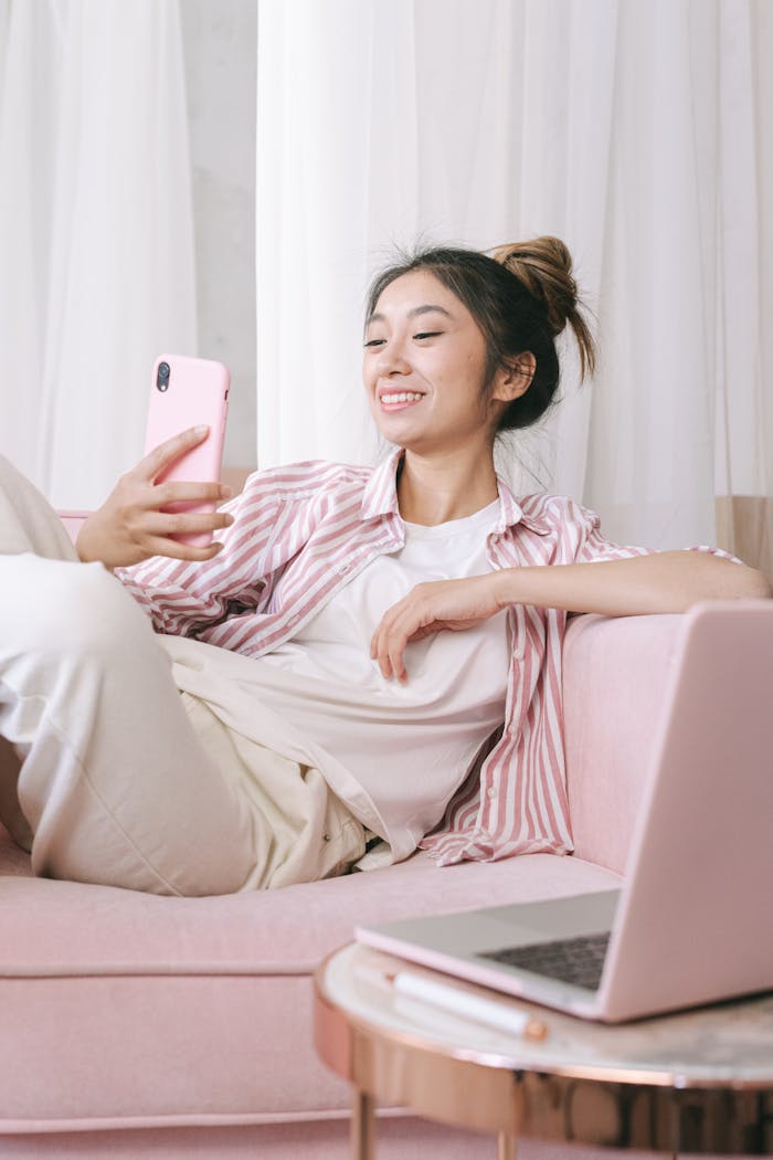 Asian woman relaxing on a pink sofa, using a smartphone and smiling at home.